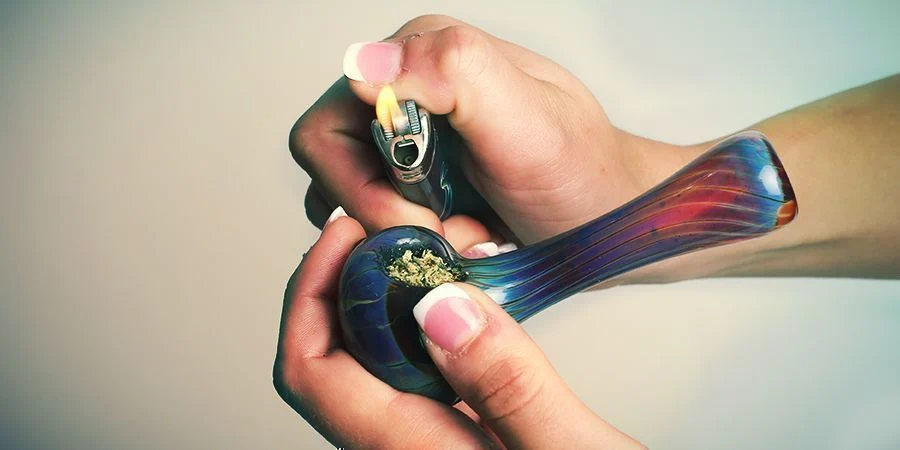 Person demonstrating how to properly pack a bowl using ground cannabis, a grinder, and a glass pipe.